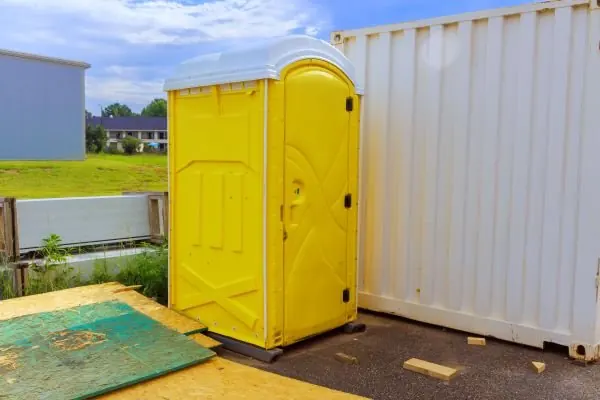 A yellow portable toilet with a white roof on plywood beside a white shipping container at a building site
