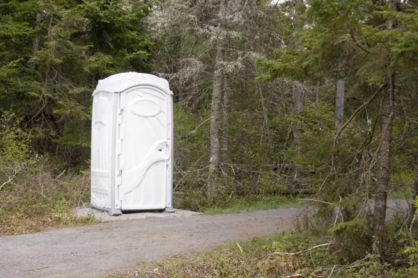 A white portable toilet on a gravel pad at the edge of a narrow road through evergreen woods